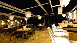 Un espace de restauration en plein air doté d’un sol en pierre, de tables et de chaises en bois, et abrité par une structure couverte ornée de lustres suspendus.