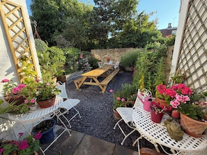 A patio with a wooden table and white metal chairs, surrounded by potted plants and a stone wall.