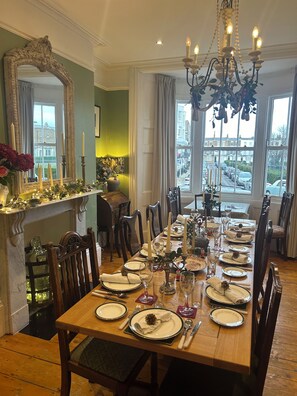 A dining room with a long table set for a meal, chairs, a chandelier, and a view of the street through large windows. - Ramsgate Retreats | Latimer House, families, big birthdays, golf, parking, 6-14 (ramsgate)
