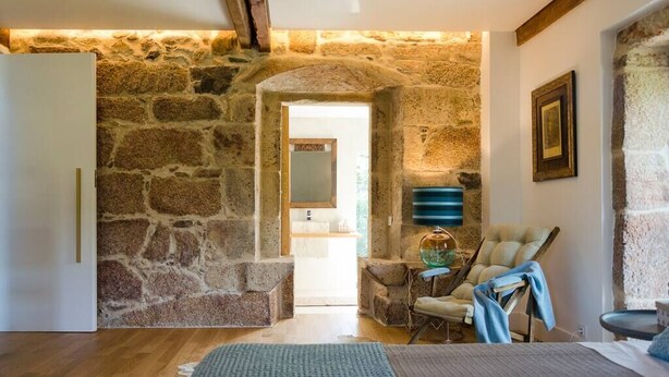 A room with stone walls, a wooden ceiling, a bed, a chair, and a table with a glass top. - A Unique Home, For A Truely Unique Experience (Mos)