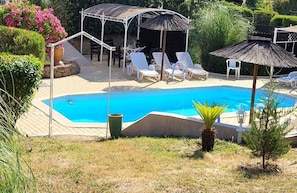 A swimming pool with lounge chairs and umbrellas, surrounded by greenery and a pergola structure.