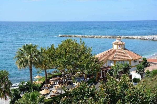 A coastal resort with a dining area, palm trees, and a view of the sea.
