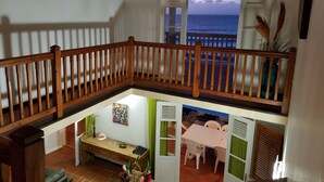 Un balcón con baranda de madera que da a un área de comedor con una mesa, sillas, un banco y vista al mar.