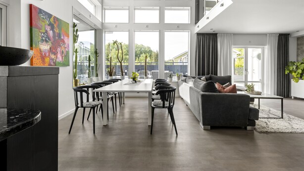 Een moderne woonkamer met een grijze bank, een eethoek met een witte tafel en grote ramen met uitzicht op groen.