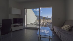 A modern living room with a glass coffee table, a sofa, and a TV. A sliding glass door leads to an outdoor terrace with a table and chairs. - Amazing apartment on the seafront in Roses Inmosantos Oasis B8 (Rosas / Roses)