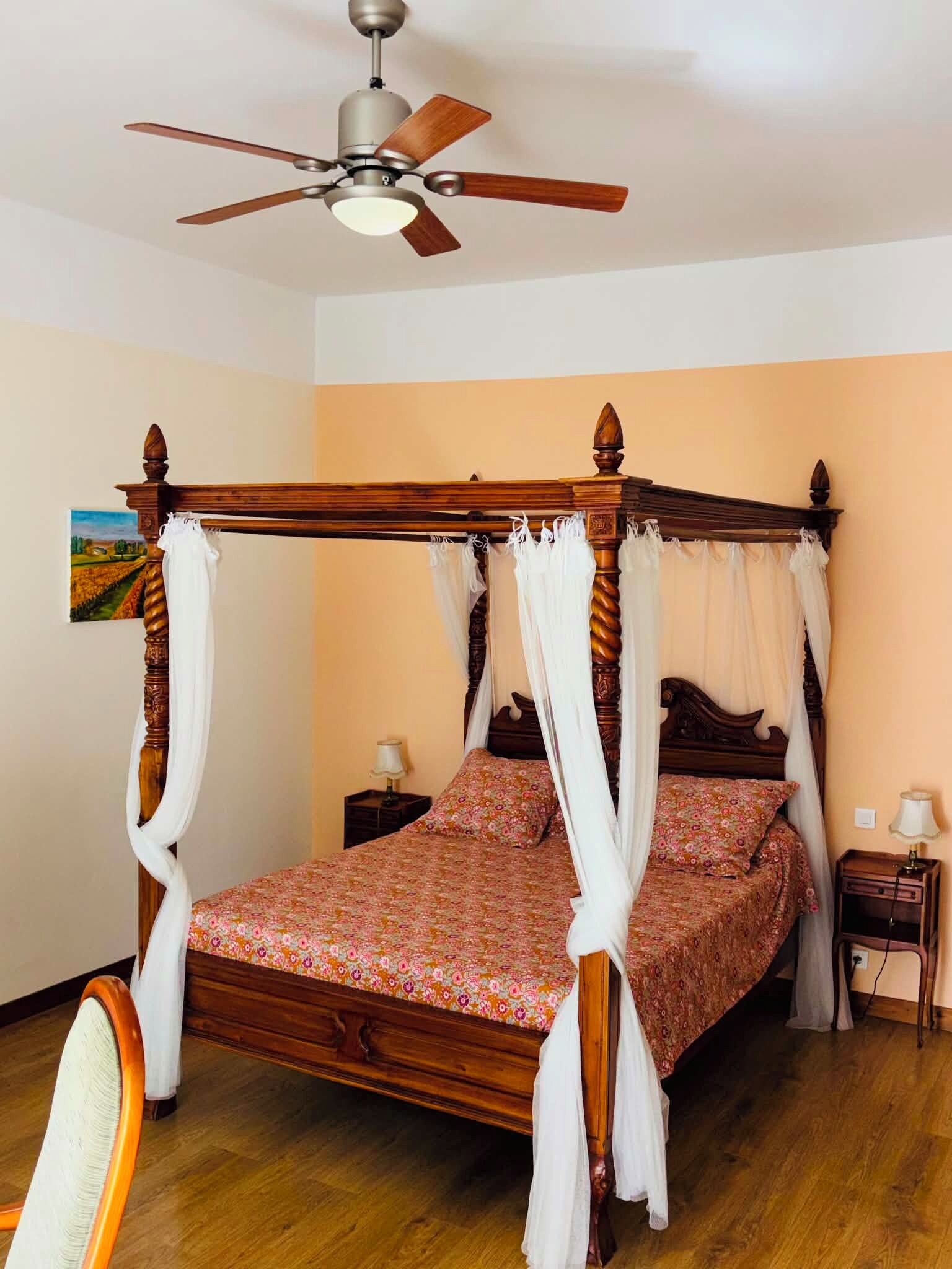 Una cama con dosel y cuatro columnas, un ventilador de techo y una mesita de noche con lámpara.