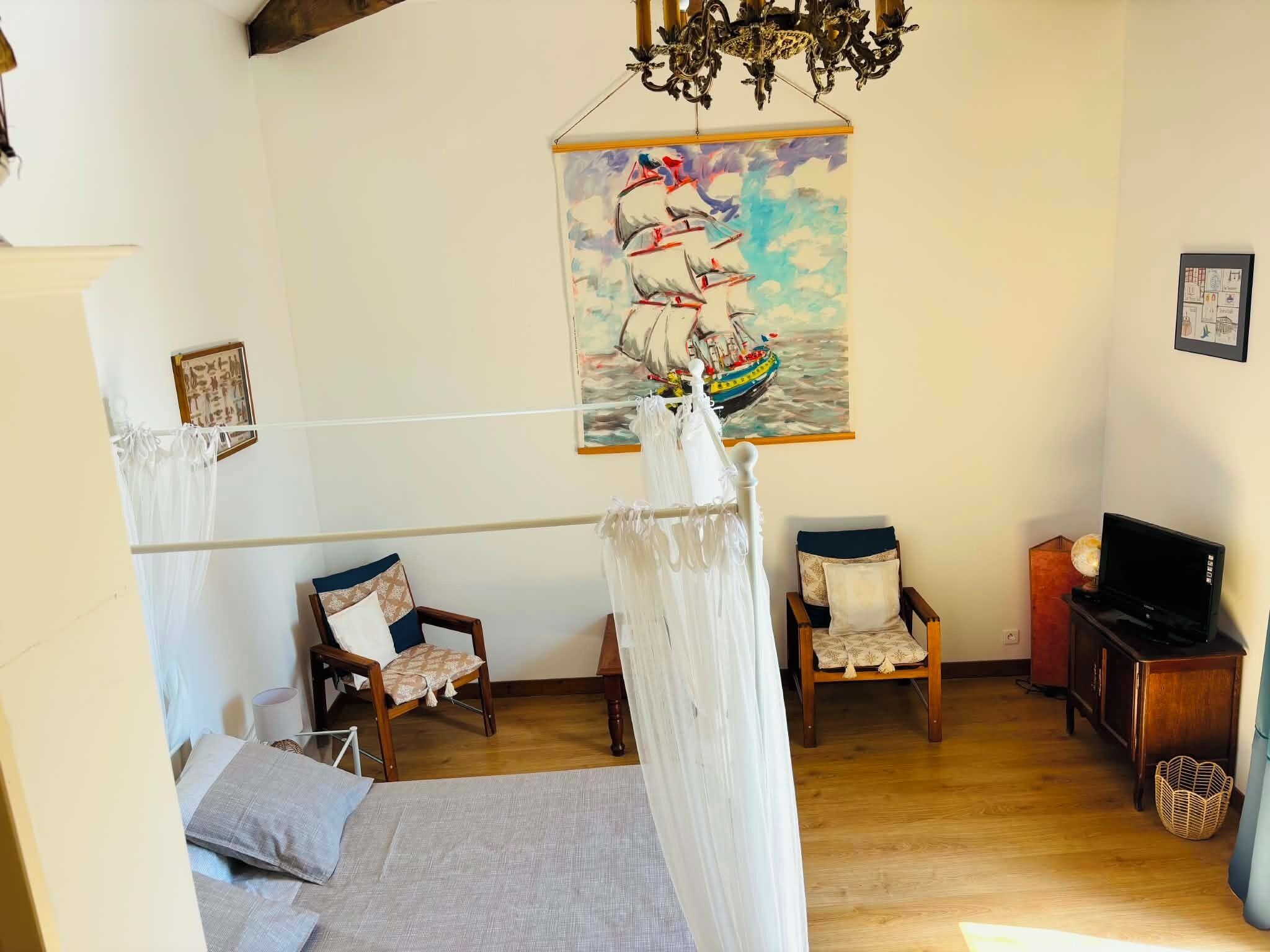 A room with a bed, two chairs, a TV, a painting of a ship, and a chandelier.