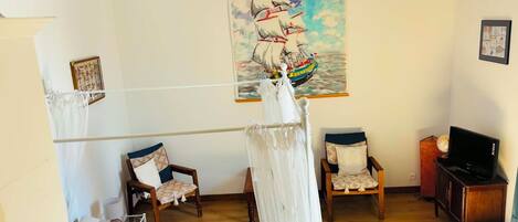 A room with a bed, two chairs, a TV, a painting of a ship, and a chandelier.