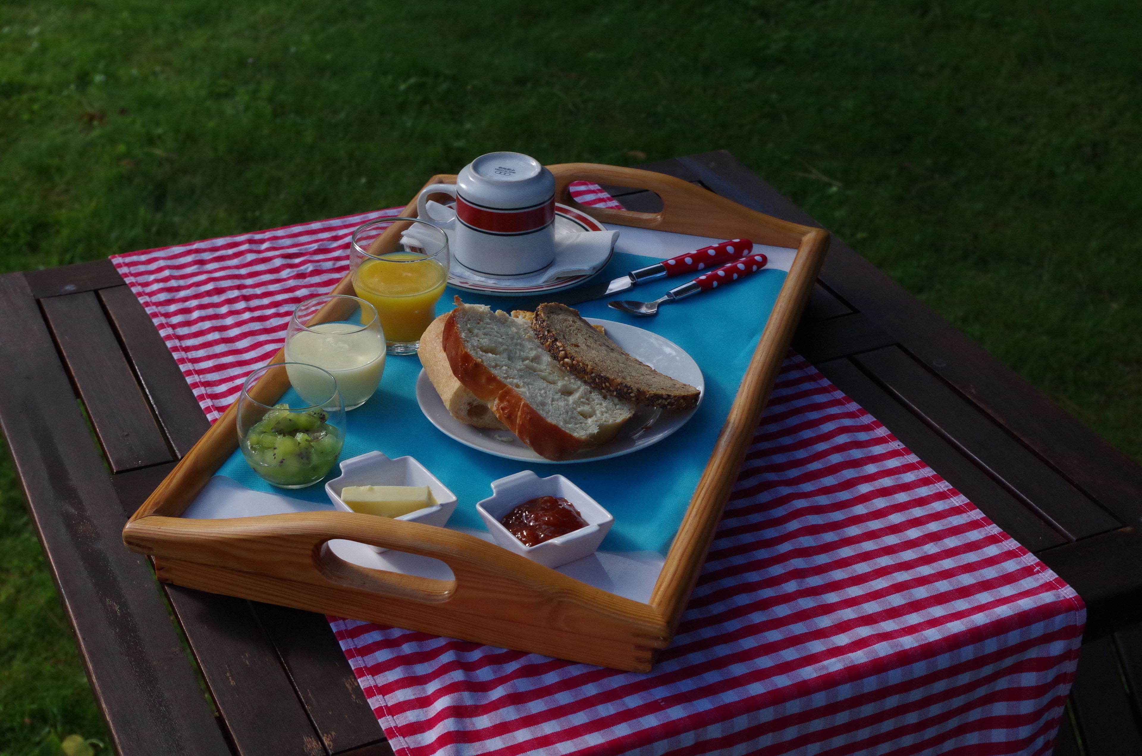 A picnic setup on a wooden table with a red and white checkered tablecloth, featuring a tray with bread, butter, jam, a glass of juice, and a cup of tea.