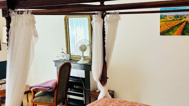 A room with a four-poster bed, a desk with a chair, a fan, and a painting on the wall. - Maison d'Hôtes Le Cadran Solaire (Courcelles)