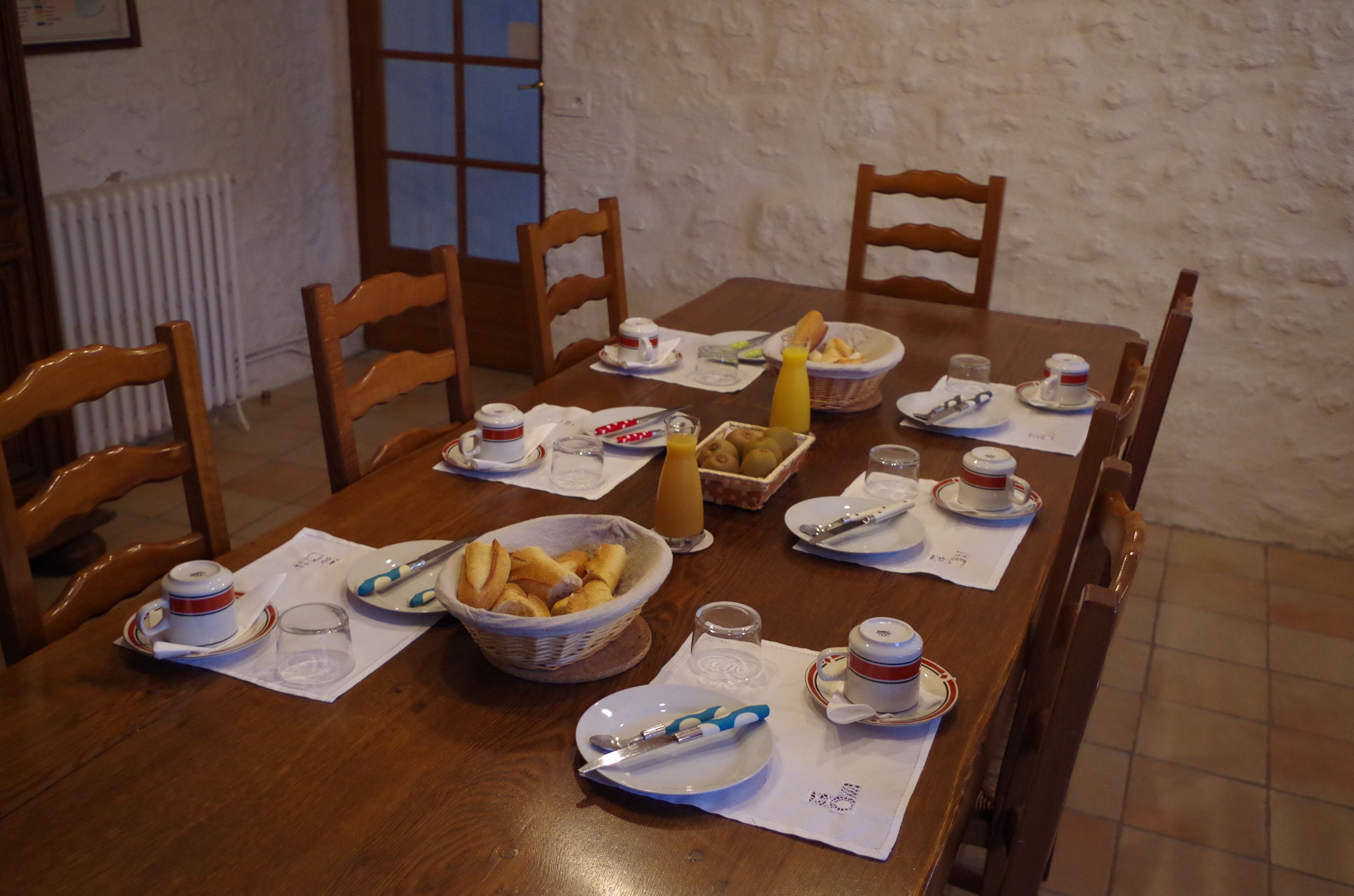 A wooden dining table set with breakfast items, including bread, fruit, and beverages, in a room with white walls and wooden chairs.