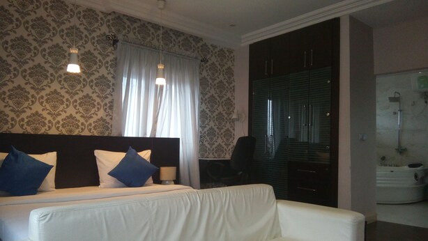 A hotel room with a bed, a white sofa, a desk, and a bathroom with a shower. - Orchid Suite & Apartment (Abuja)