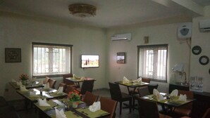 A dining area with multiple tables set for two, each with a green placemat, white napkins, and cutlery. There are two windows with bars, and an air conditioning unit mounted on the wall. - Orchid Suite & Apartment (Abuja)