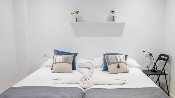 A hotel room with a bed, two pillows, a heart-shaped towel arrangement, a small shelf with flowers, and a chair. - Apartamento El Patio de Cánovas (Cádiz)