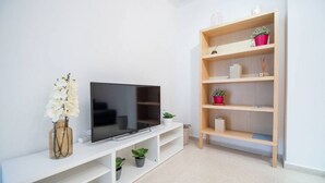 A modern living room with a flat-screen TV on a white entertainment unit, a wooden bookshelf with various items, and a vase with white flowers. - Apartamento El Patio de Cánovas (Cádiz)