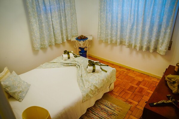 A small, neatly arranged bedroom with a single bed, a small table, and a chair. There are floral curtains on the windows, and a small decorative item is placed on the windowsill.