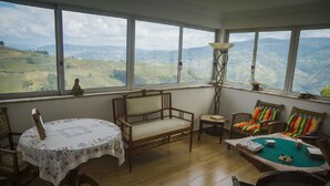 Una habitación con un ventanal grande que ofrece vistas de colinas ondulantes. Se observa una silla de madera, una mesita con mantel floreado y una silla colorida con cojines a rayas.