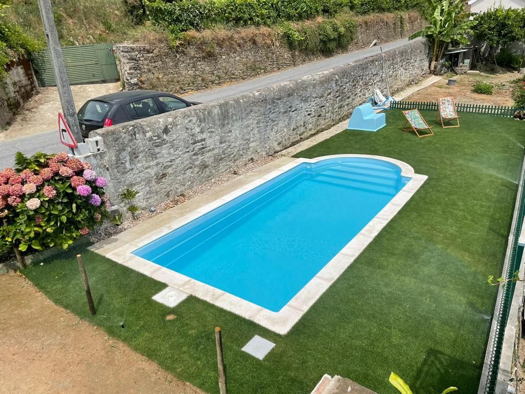 Una piscina rectangular con un área verde circundante, un muro de piedra y una carretera con un coche pasando.