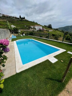 A rectangular swimming pool with a white border, surrounded by well-maintained grass, and a backdrop of a hilly landscape with buildings.