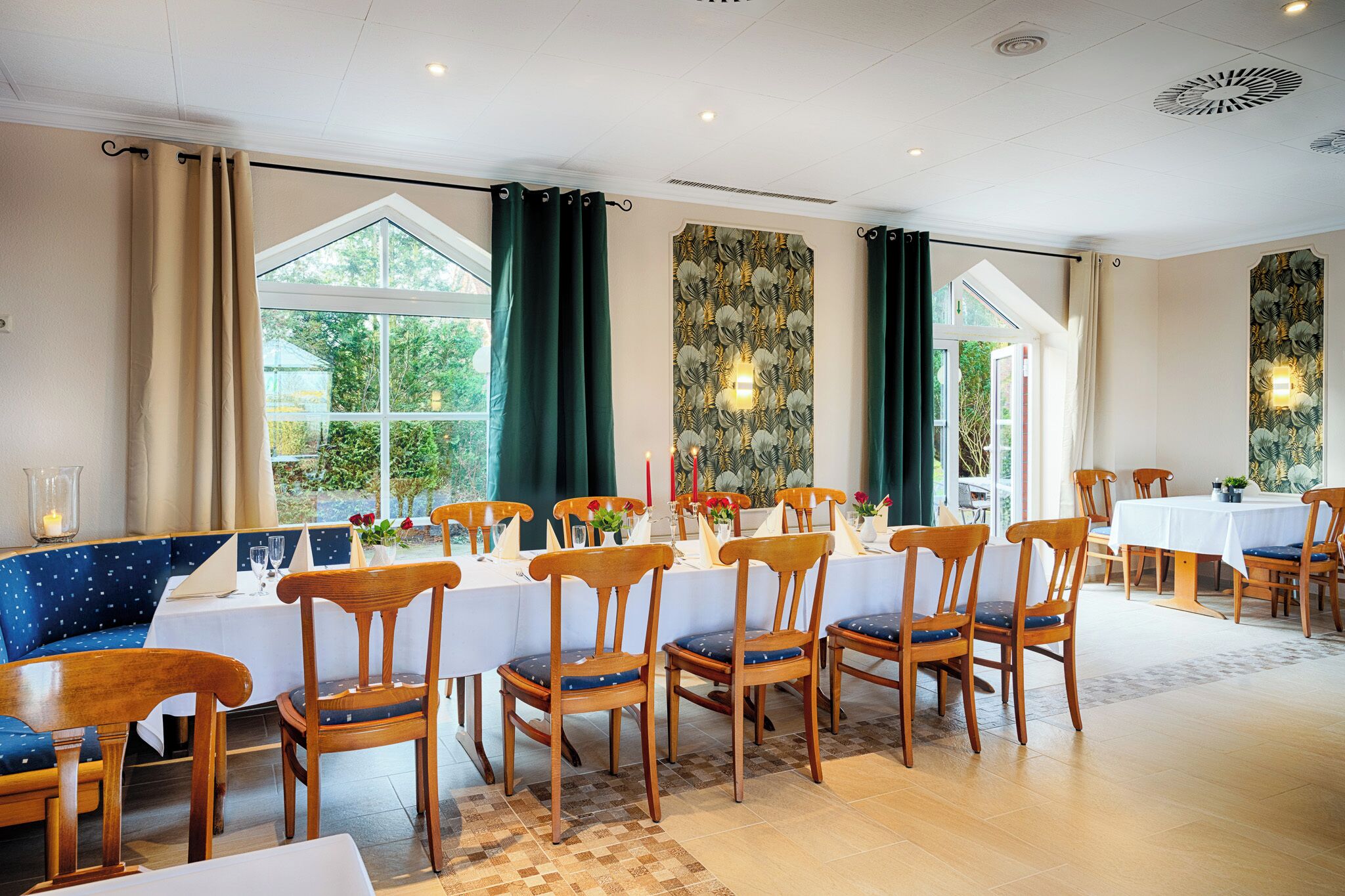 Un espace repas avec des chaises et des tables en bois, de grandes fenêtres avec des rideaux et une vue sur la verdure à l’extérieur.