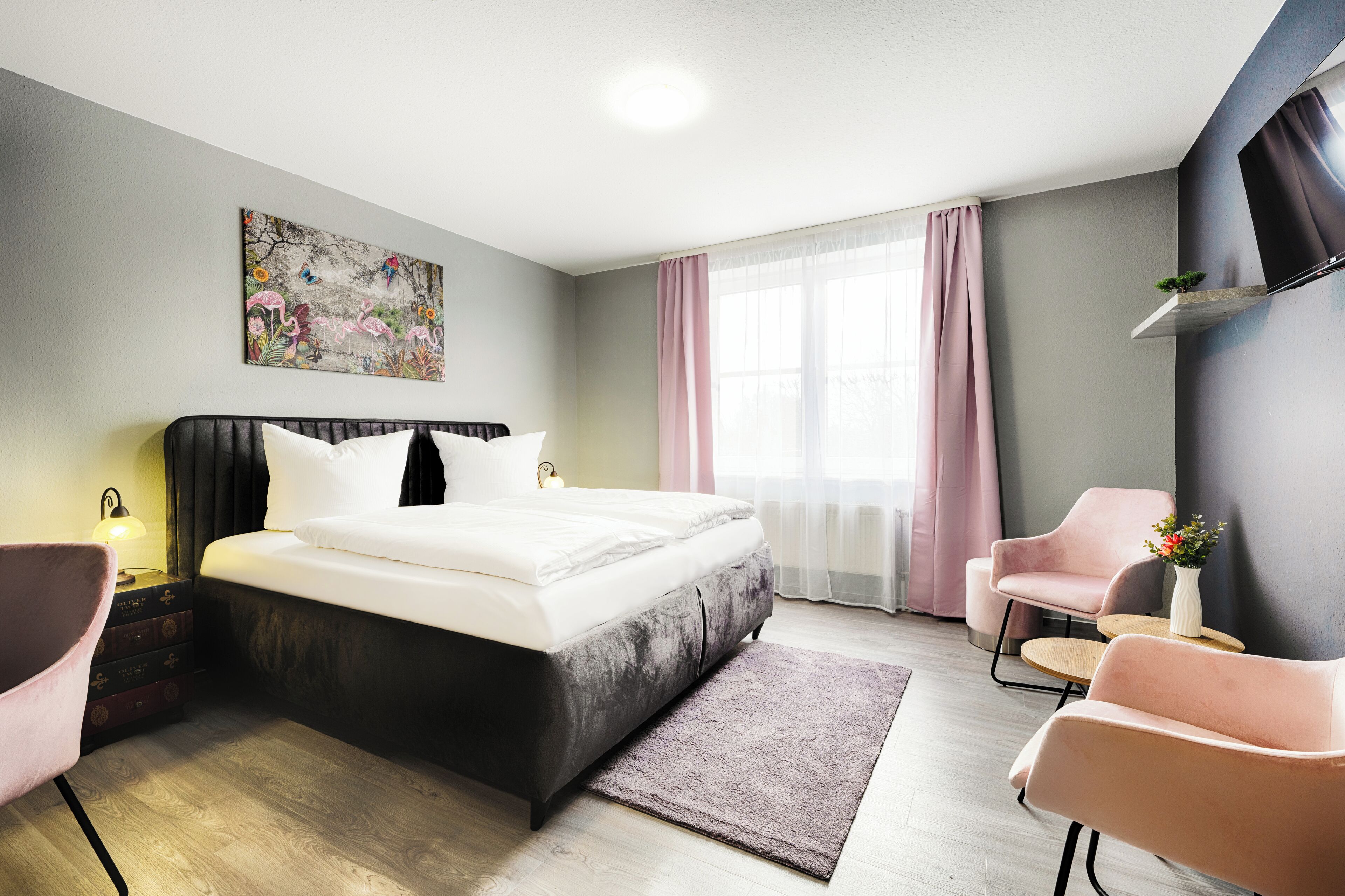 Um quarto moderno com uma cama grande, uma mesinha de cabeceira com abajur, uma cadeira, uma pequena mesa com um vaso e um quadro na parede.