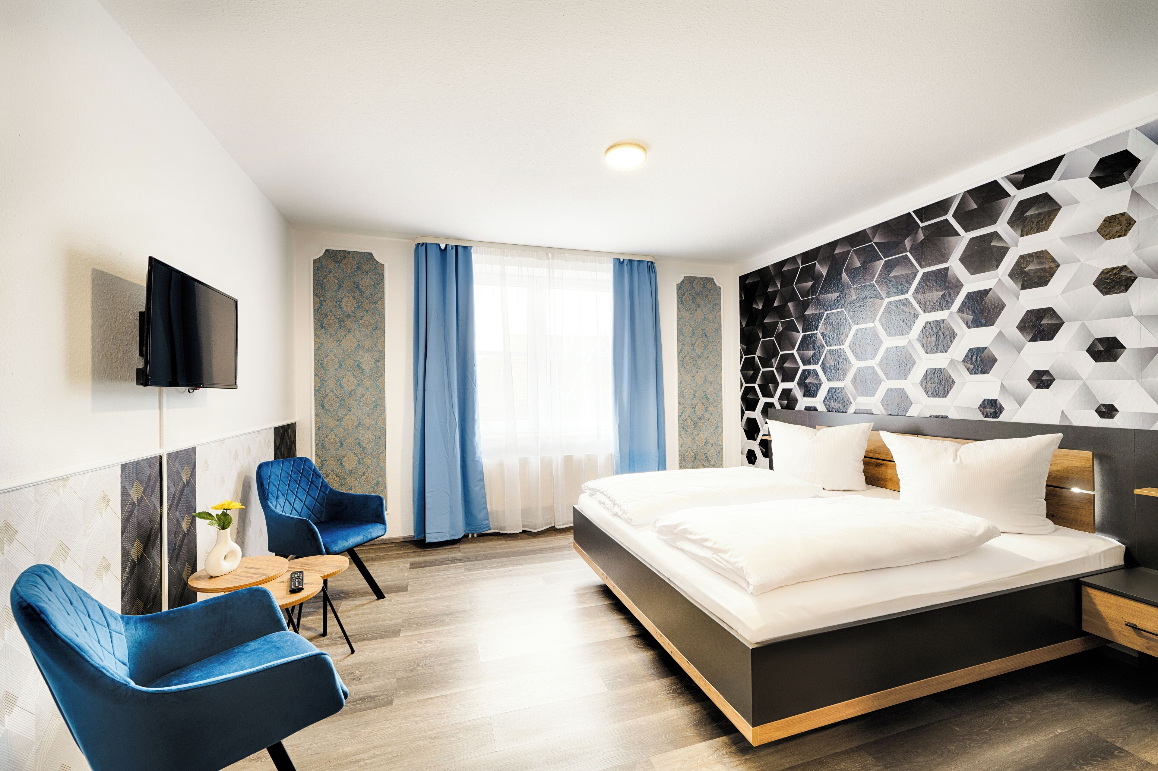 A modern hotel room with a large bed, a flat-screen TV, two blue armchairs, a small table with a vase, and a decorative wall panel.