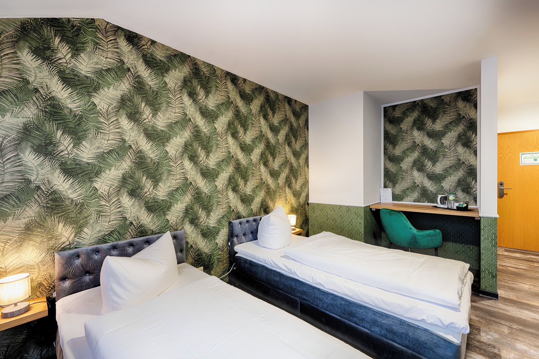 A hotel room with two beds, a green chair, a desk with a mirror, and a patterned wall.