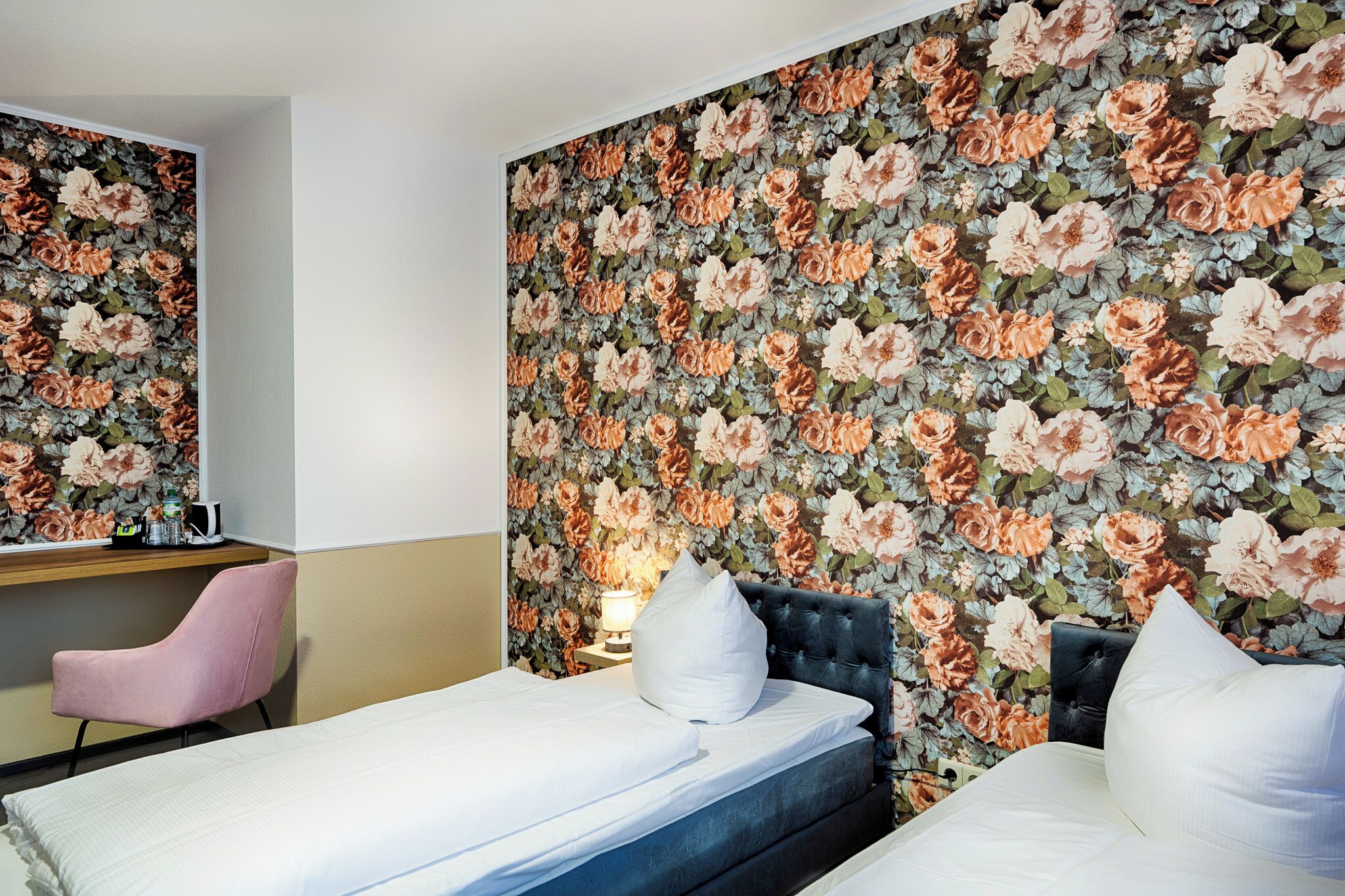 A hotel room with a floral wallpaper, a bed, a chair, and a bedside table with a lamp.