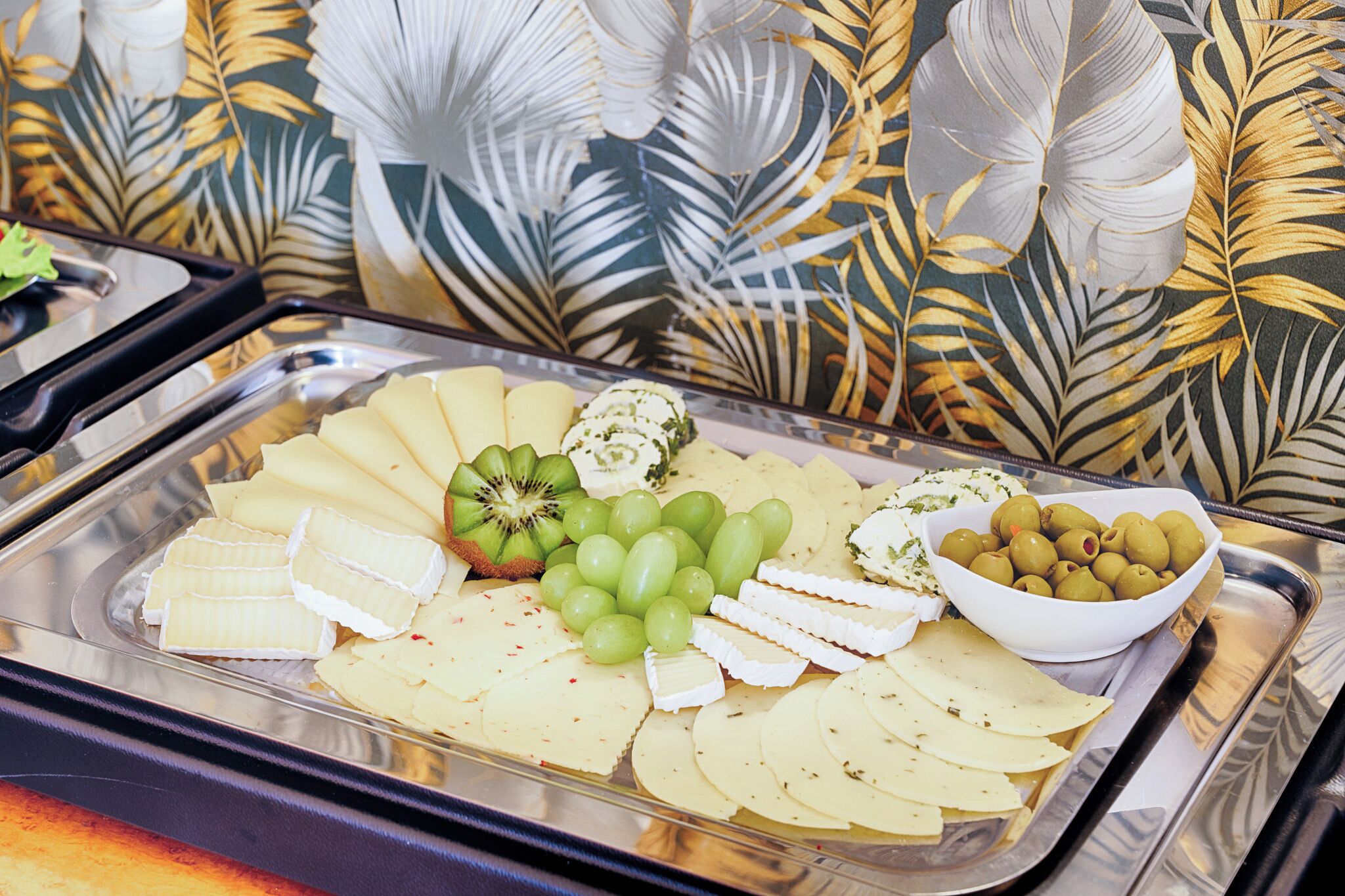 A buffet tray with various cheeses, grapes, kiwi, and olives.