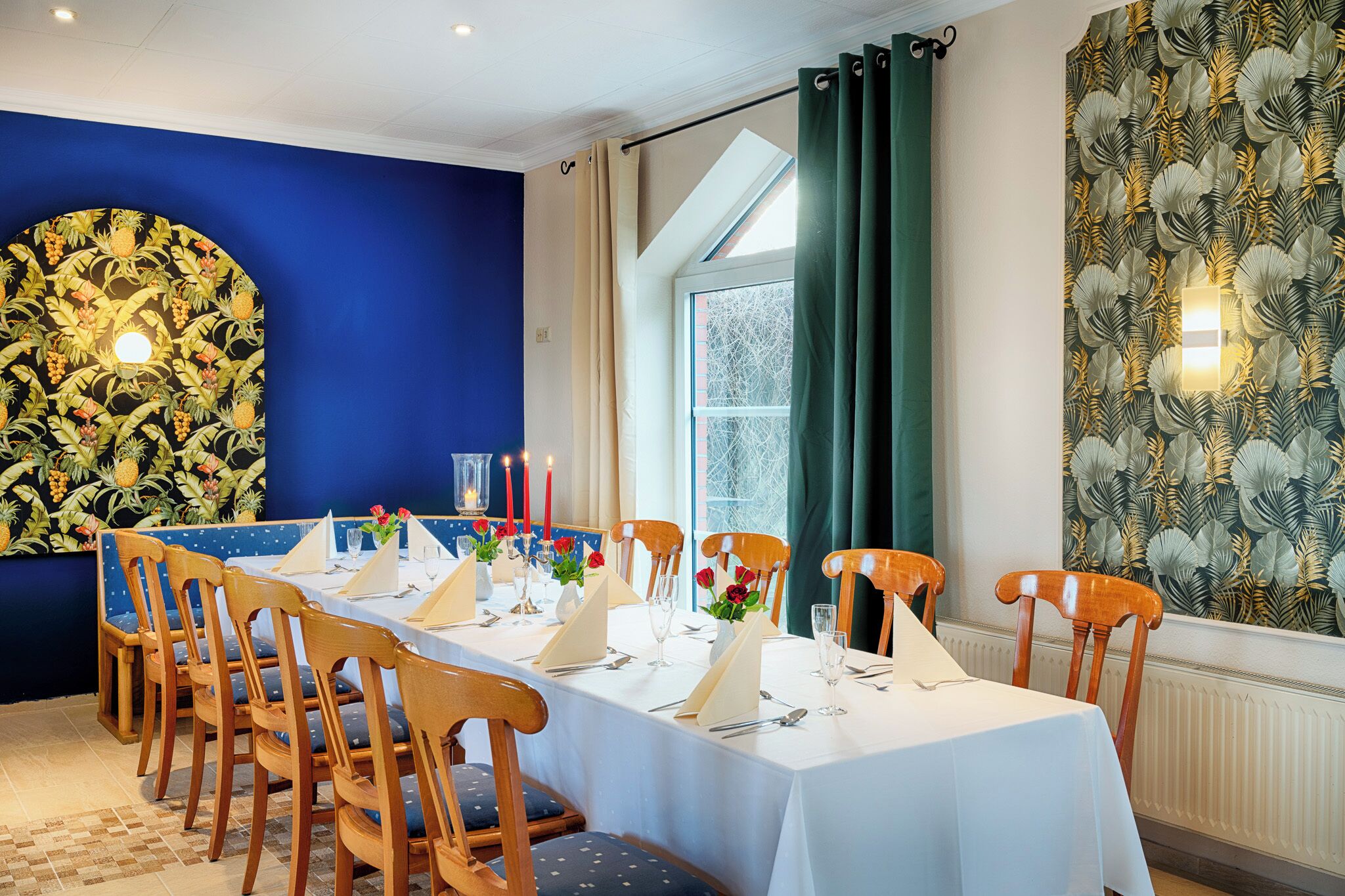 Een eethoek met een tafel voor zes personen, houten stoelen en een blauwe wand met tropische behang.