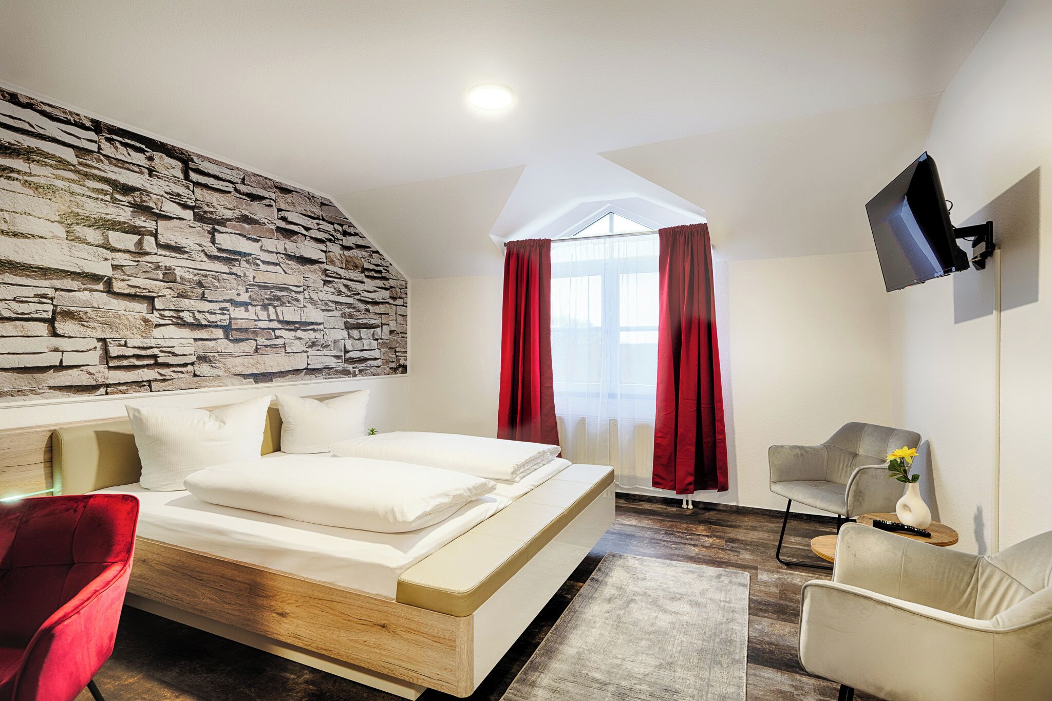 A modern bedroom with a stone wall, a large bed, red curtains, a flat-screen TV, and a seating area with a vase of flowers.