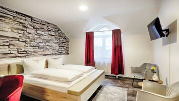 A modern bedroom with a stone wall, a large bed, red curtains, a flat-screen TV, and a seating area with a vase of flowers.