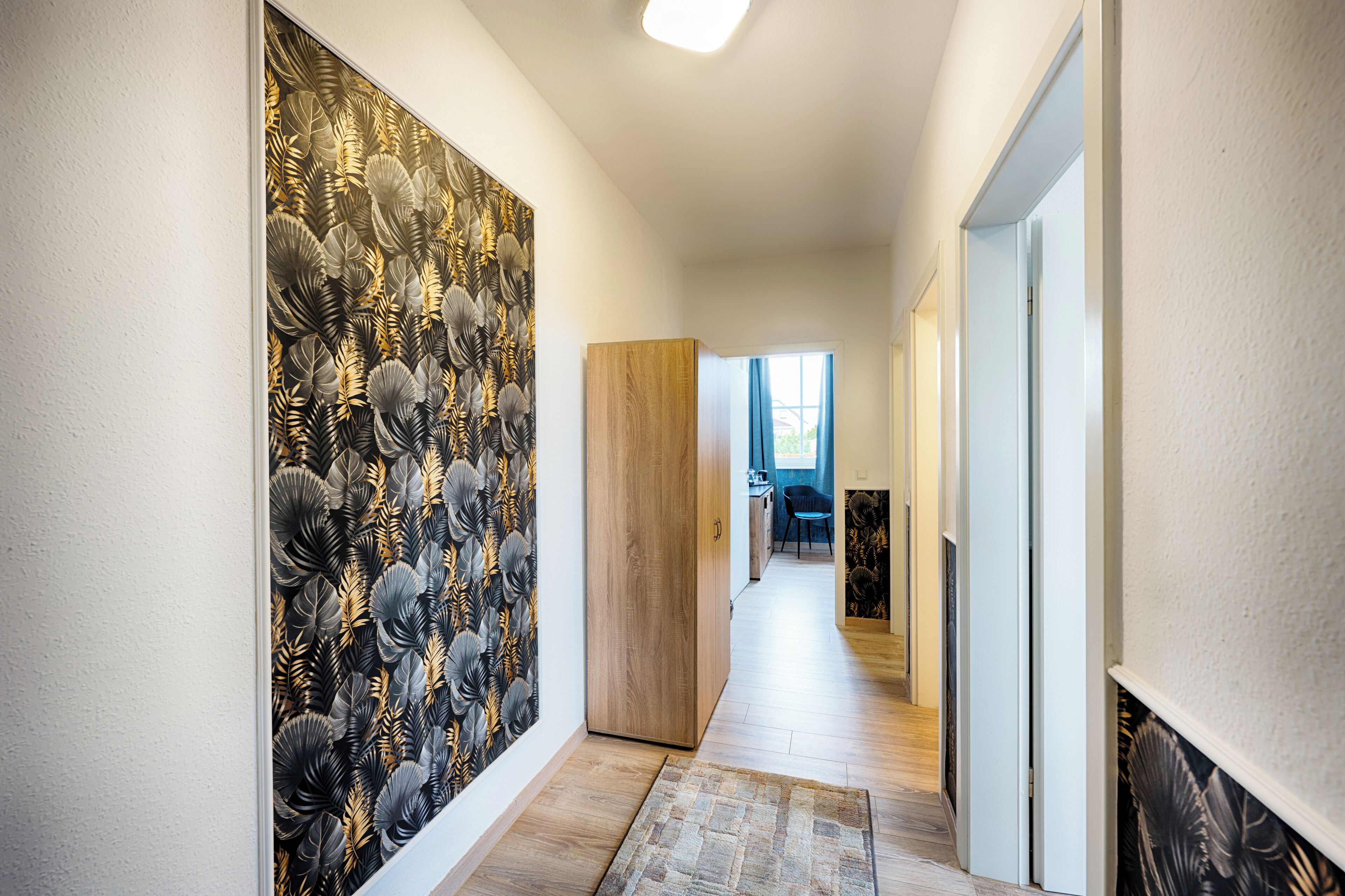 A hallway with a patterned wallpaper, a wooden door, and a rug leading to a room with a blue chair and a desk.