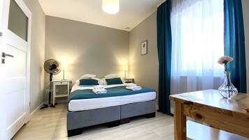 Ein Hotelzimmer mit Bett, Nachttischen, einem Schreibtisch, einem Sessel, einem Ventilator, einem Fenster mit Vorhängen und einer Vase mit Blumen.