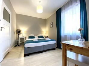 Une chambre d’hôtel comprenant un lit, des tables de chevet, un bureau, une chaise, un ventilateur, une fenêtre avec des rideaux et un vase avec des fleurs.