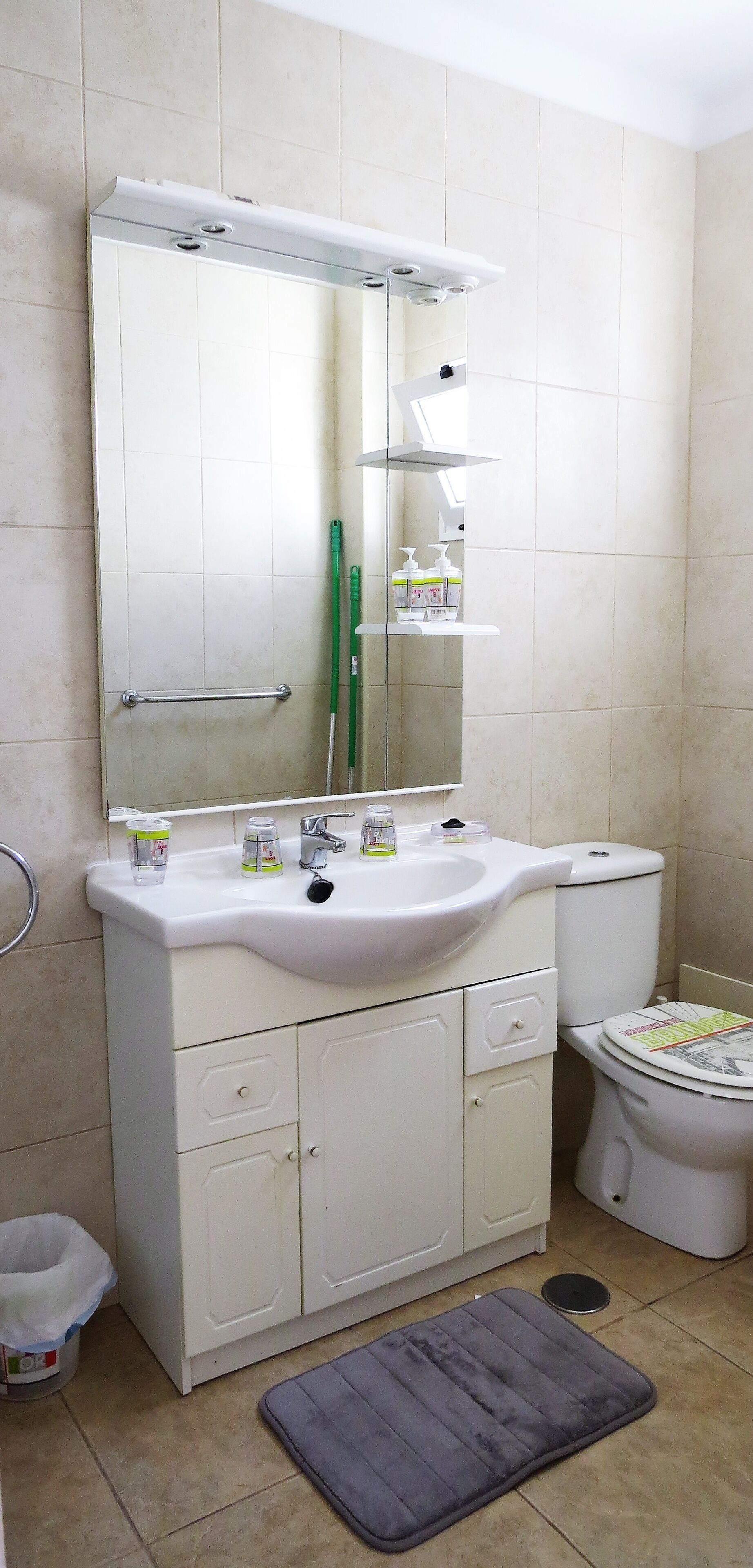 Ein Badezimmer mit einem weißen Waschbecken, einem Spiegel und einer Toilette. Auf dem Waschbecken liegen Toilettenartikel und auf dem Boden liegt eine Badmatte.