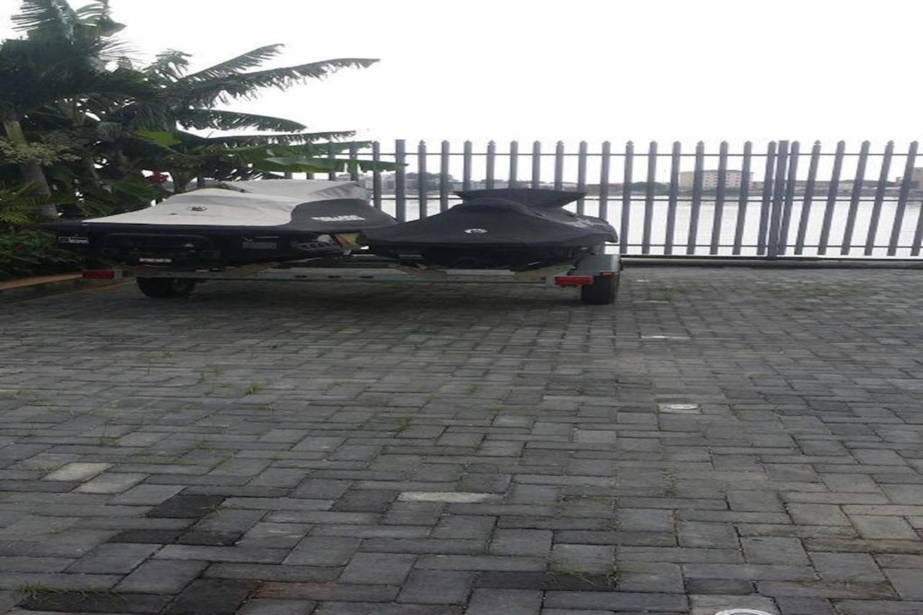 Un jet ski est posé sur une remorque, le tout étant stationné sur une surface goudronnée, avec une clôture métallique et des palmiers en arrière-plan.