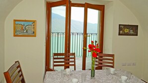 A dining area with a table set for two, a vase with red flowers, and a view of mountains through a balcony door.