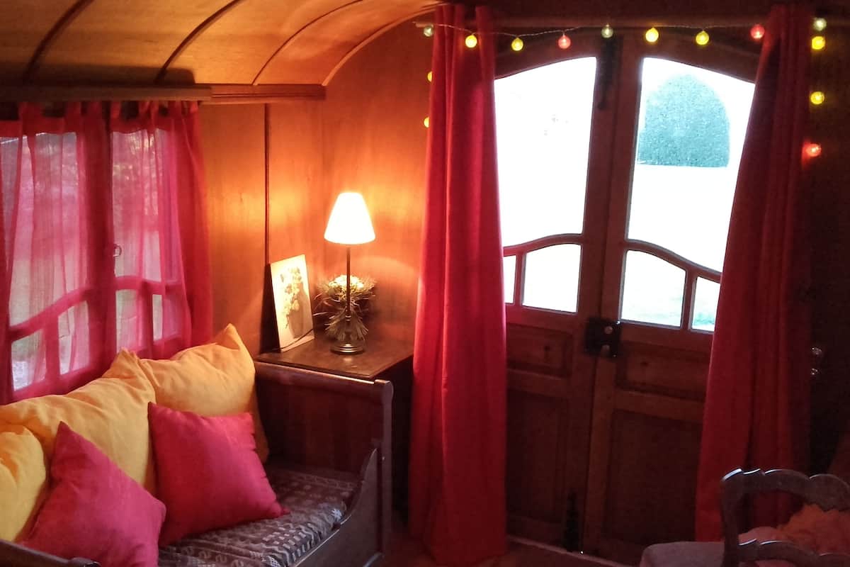 A cozy room with a wooden bench, red curtains, a table with a lamp, and a view through a window.