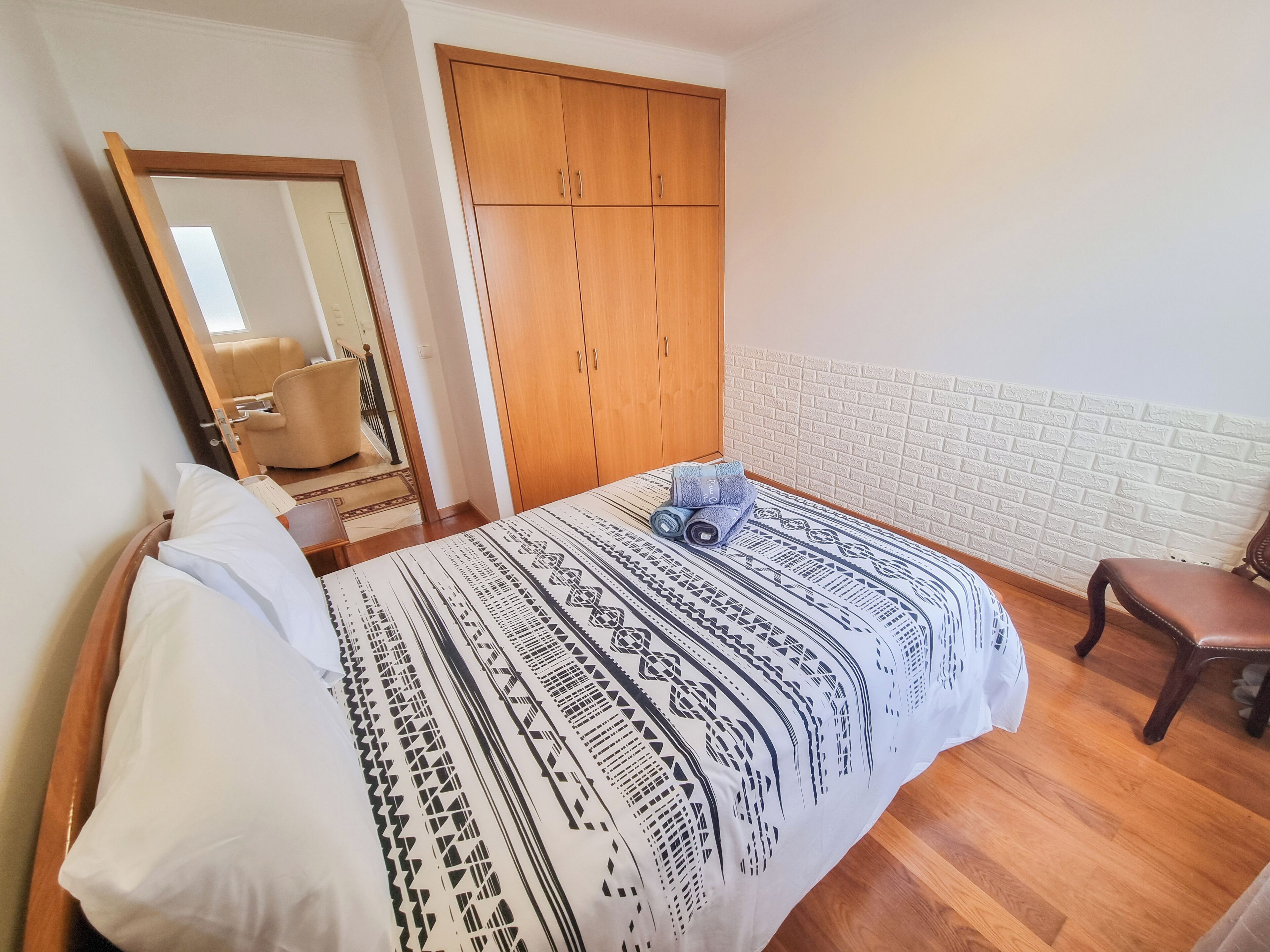 A bedroom with a bed, a wooden wardrobe, a chair, and a mirror.