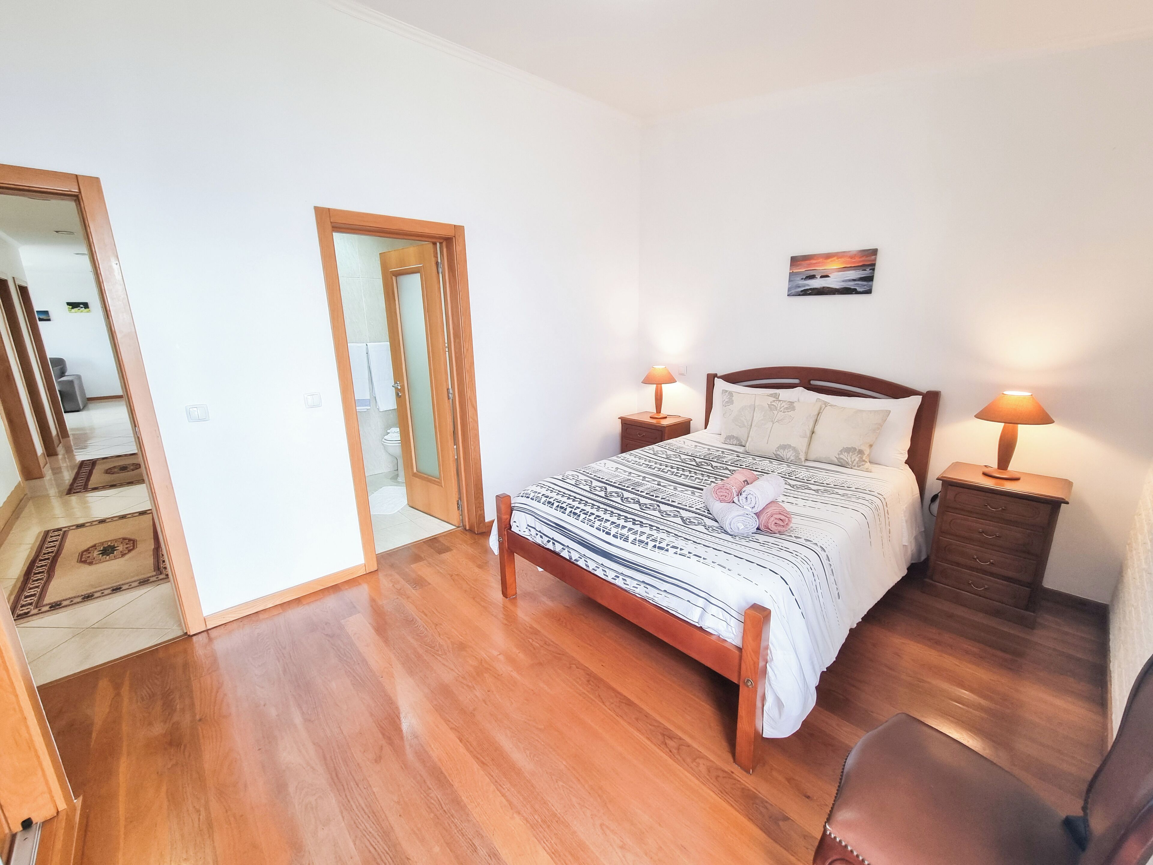 A bedroom with a wooden bed, two bedside tables with lamps, a small chair, and a view of the bathroom through an open door.