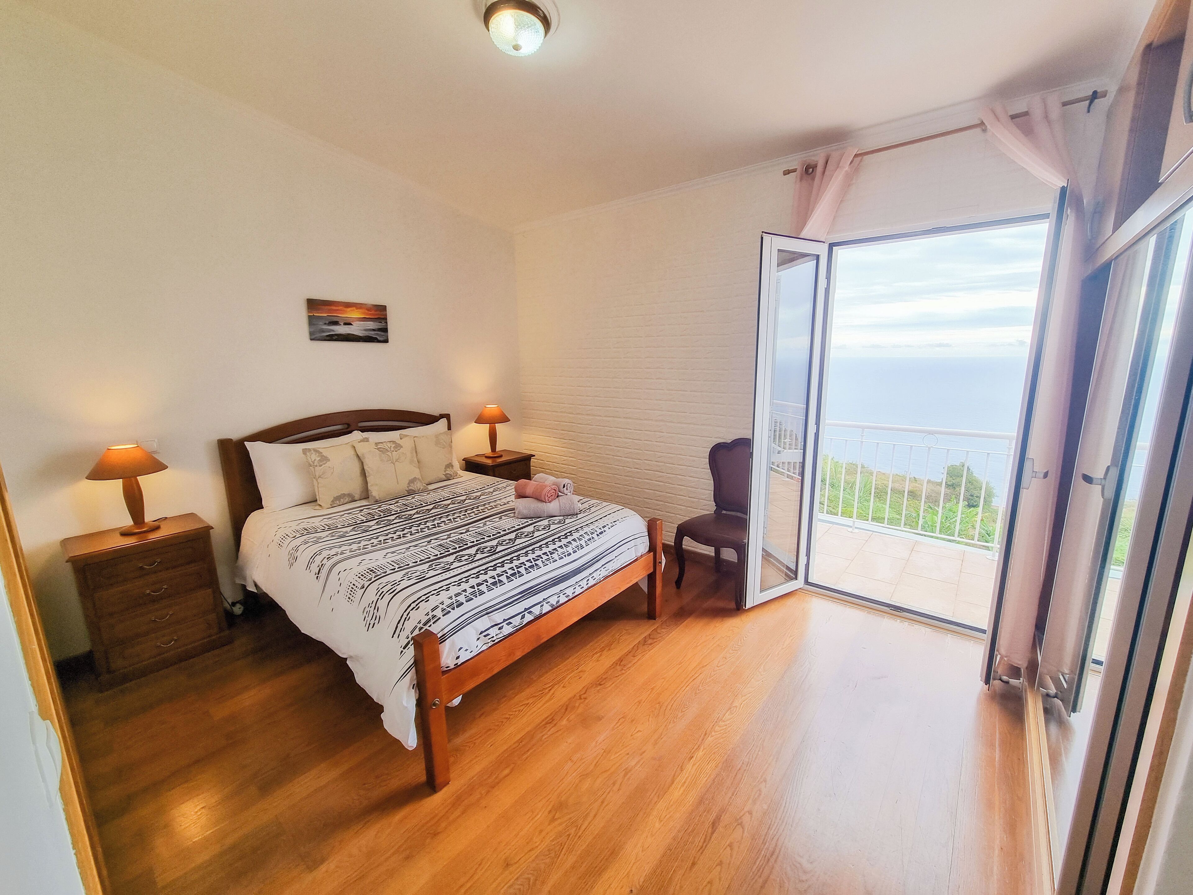 Een slaapkamer met een bed, twee nachtkastjes, een stoel, een schilderij aan de muur en een balkon met uitzicht.