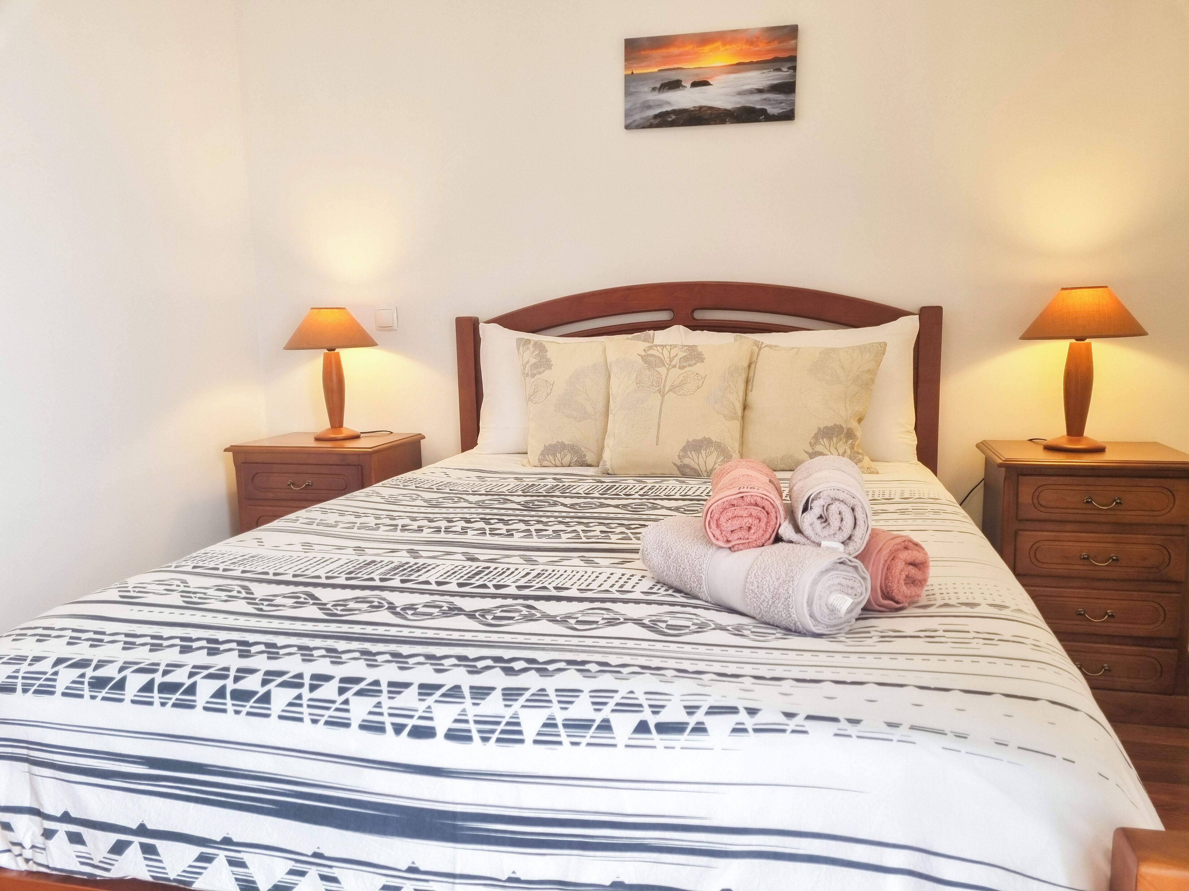 Una cama bien hecha con ropa de cama estampada, dos mesitas de noche con lámparas, y un cuadro en la pared.