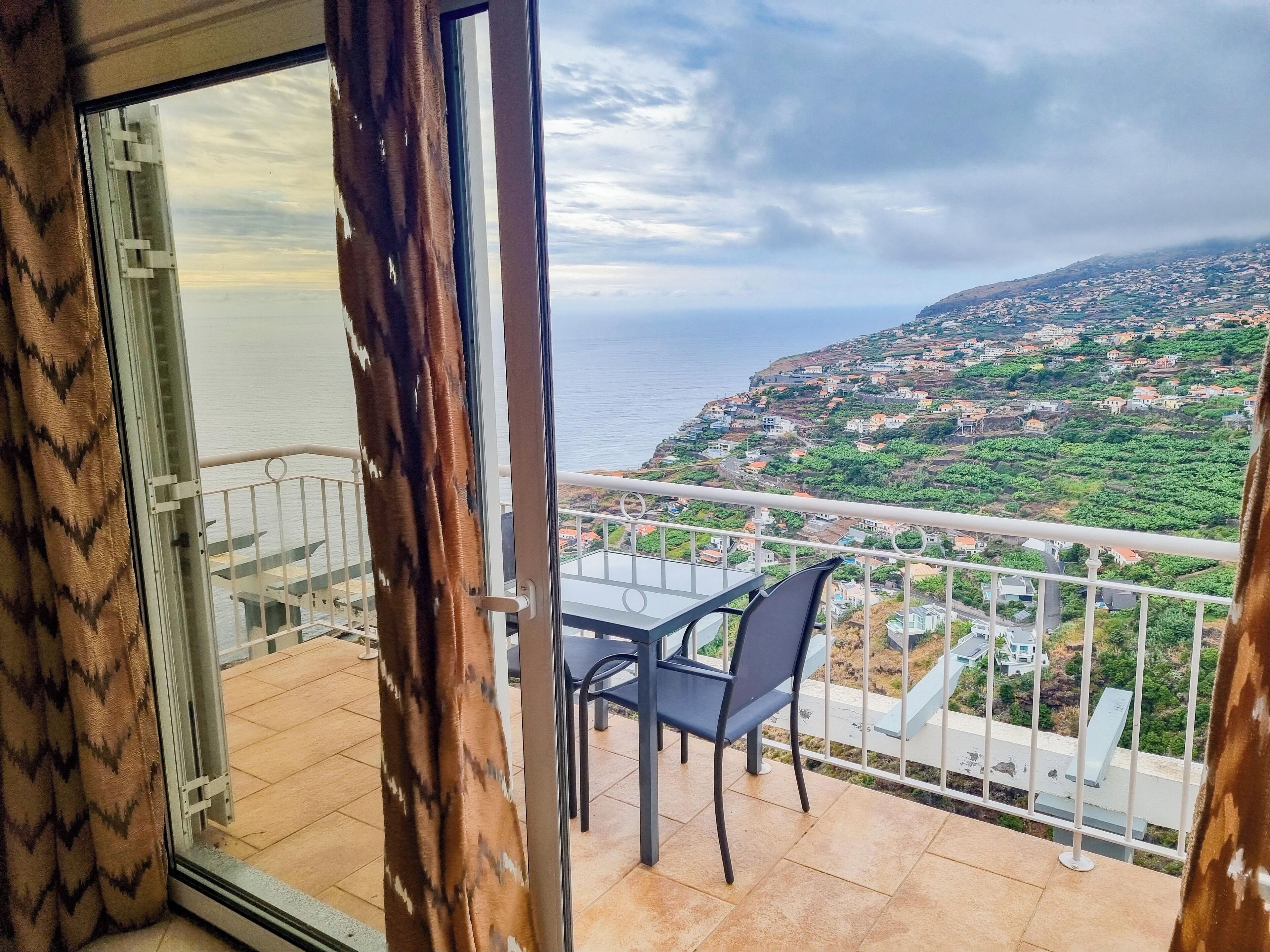 A balcony with a table and chairs, overlooking a coastal town with houses and greenery.