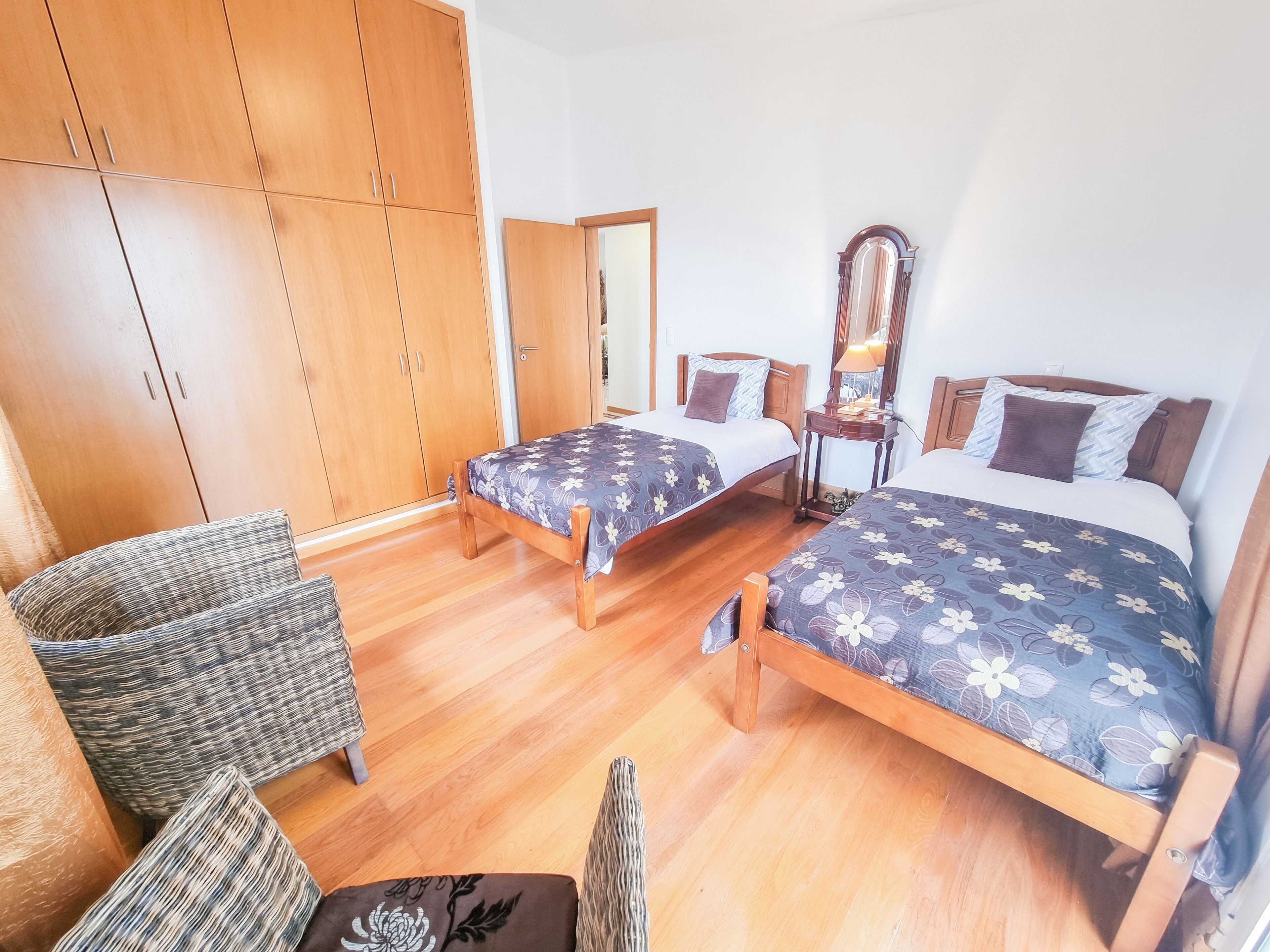 Un dormitorio con dos camas, un armario de madera, sillas de mimbre y un espejo.