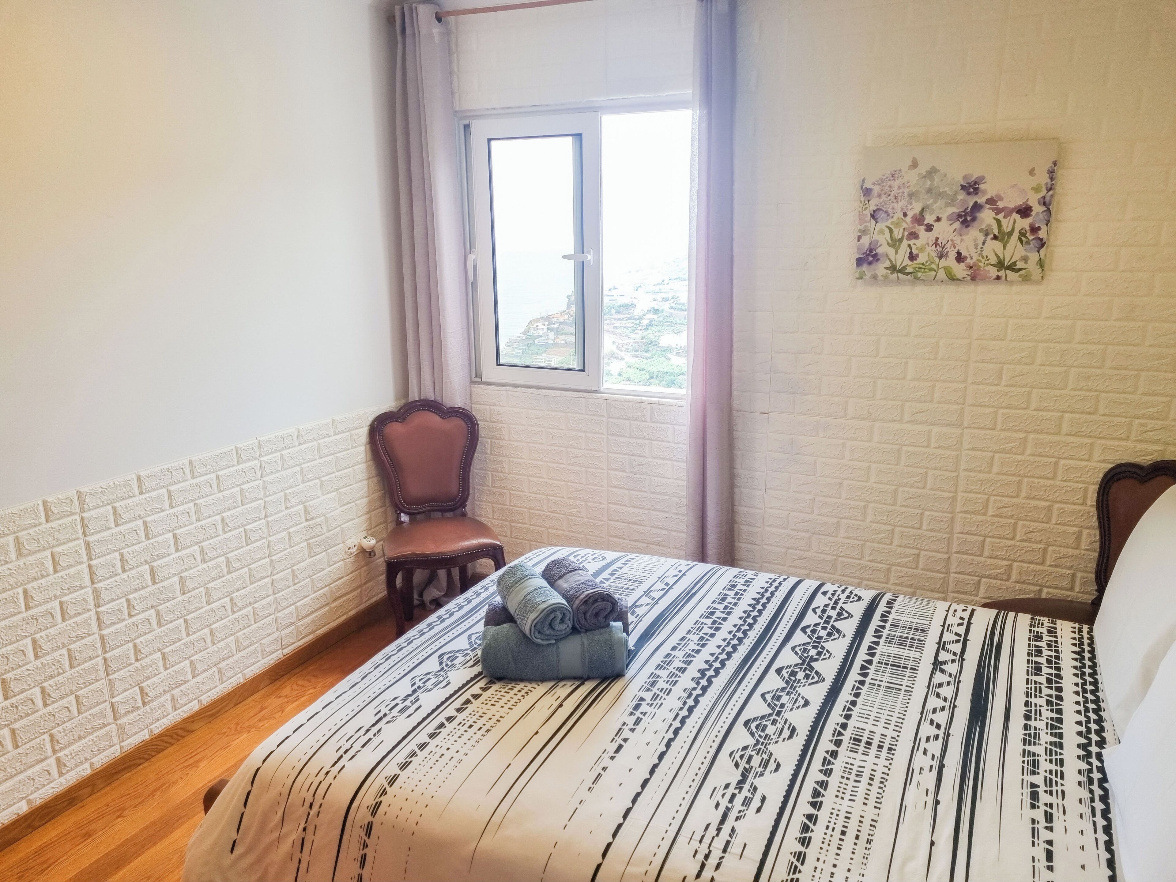 A bedroom with a patterned bedspread, a chair, a window with curtains, and a floral artwork on the wall.