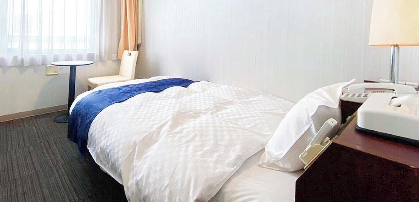 A hotel room with a bed, a chair, a small table, a lamp, and a telephone.