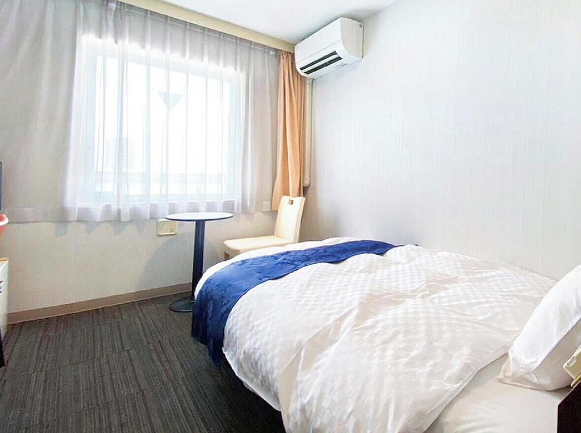 Quarto de hotel com cama, poltrona, mesinha, ar condicionado e janela com cortinas.