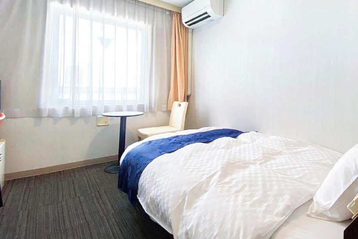 A hotel room with a bed, a chair, a small table, an air conditioning unit, and a window with curtains.