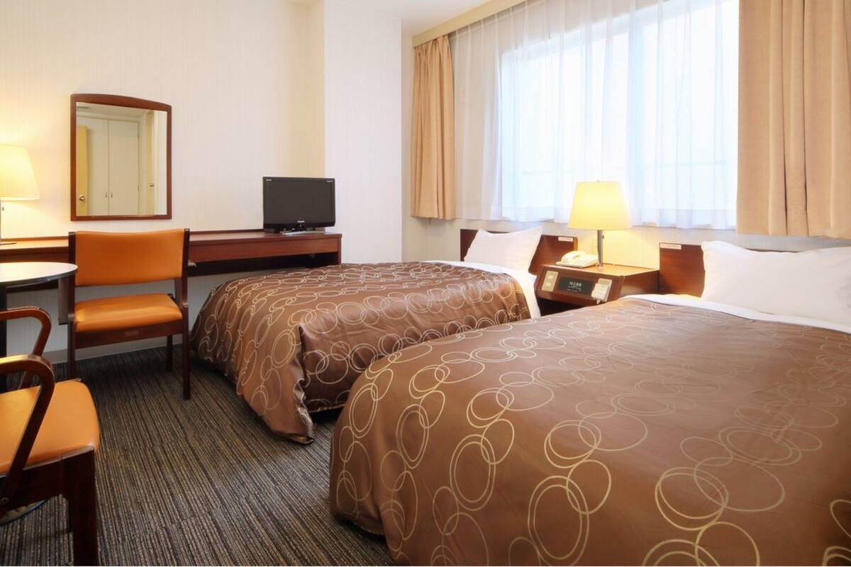 Ein Hotelzimmer mit zwei Betten, einem Schreibtisch mit Fernseher, einem Sessel und einem Fenster mit Vorhängen.